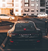 A luxury sports car is parked in a residential area, surrounded by modern apartment buildings. The sleek vehicle is prominently displayed, catching the warm glow of the setting sun. Nearby, other cars are parked, hinting at an urban environment.