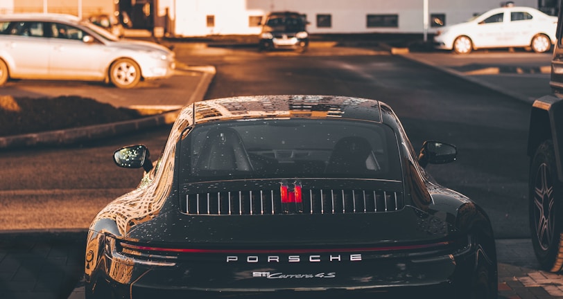A luxury sports car is parked in a residential area, surrounded by modern apartment buildings. The sleek vehicle is prominently displayed, catching the warm glow of the setting sun. Nearby, other cars are parked, hinting at an urban environment.