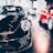 A sleek black sports car is prominently displayed indoors, surrounded by other luxury vehicles. The environment suggests a high-end showroom or dealership with bright lighting and modern architecture. The flooring is reflective, adding to the luxurious tone.