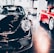 A sleek black sports car is prominently displayed indoors, surrounded by other luxury vehicles. The environment suggests a high-end showroom or dealership with bright lighting and modern architecture. The flooring is reflective, adding to the luxurious tone.