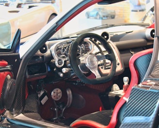 A luxury sports car interior featuring a sleek, high-tech dashboard with a carbon fiber finish. The steering wheel is prominently displayed, surrounded by a variety of meticulously designed control panels. Red accents highlight the seats and parts of the door, contributing to the contemporary design aesthetic.