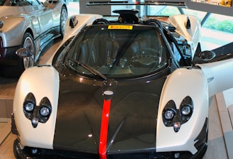 A luxury sports car is displayed in a showroom with its doors open. The car features a sleek design with a black and white color scheme, accented by a bold red stripe down the center of the front. The interior of the showroom includes other high-end cars and various automotive displays and memorabilia on the walls and shelves.