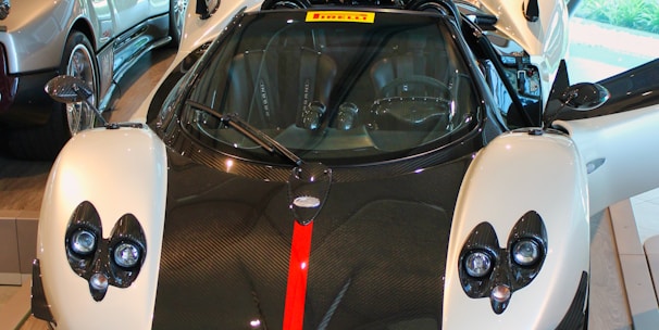 A luxury sports car is displayed in a showroom with its doors open. The car features a sleek design with a black and white color scheme, accented by a bold red stripe down the center of the front. The interior of the showroom includes other high-end cars and various automotive displays and memorabilia on the walls and shelves.