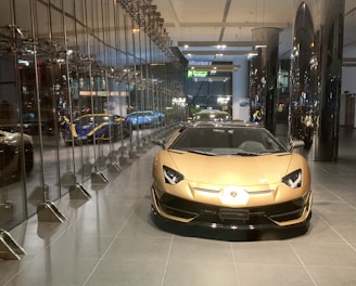 A luxurious sports car showroom with a prominent gold-colored car in the foreground. The showroom features reflective glass walls and a sleek, modern design. Several other high-end cars can be seen in the background, including a blue and yellow car. The showroom floor is clean and polished, and modern lighting fixtures enhance the upscale atmosphere of the space.