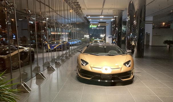 A luxurious sports car showroom with a prominent gold-colored car in the foreground. The showroom features reflective glass walls and a sleek, modern design. Several other high-end cars can be seen in the background, including a blue and yellow car. The showroom floor is clean and polished, and modern lighting fixtures enhance the upscale atmosphere of the space.