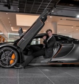 A luxury sports car with its driver-side door open, revealing a man in a suit seated inside. The sleek vehicle features a streamlined design with orange accents on the wheels and body. The setting appears to be a modern showroom with overhead lighting and reflective surfaces.