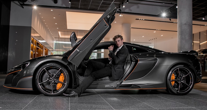A luxury sports car with its driver-side door open, revealing a man in a suit seated inside. The sleek vehicle features a streamlined design with orange accents on the wheels and body. The setting appears to be a modern showroom with overhead lighting and reflective surfaces.