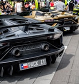 A row of luxury sports cars is parked on a street, with people gathered around them. The cars have sleek, aerodynamic designs, and the one in the foreground has a customized license plate. The scene is set against a backdrop of trees and a bright, sunny atmosphere.