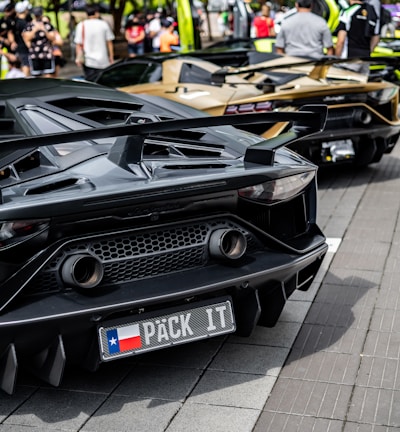 A row of luxury sports cars is parked on a street, with people gathered around them. The cars have sleek, aerodynamic designs, and the one in the foreground has a customized license plate. The scene is set against a backdrop of trees and a bright, sunny atmosphere.
