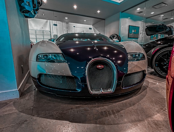 A luxury sports car is parked indoors under a ceiling with recessed lighting. The sleek and reflective surfaces of the car reflect the lights above. The room has mirrors on the walls, enhancing the sense of space and adding to the modern and sophisticated setting.