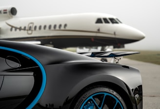 A luxury sports car with sleek black bodywork and blue accents is prominently featured in the foreground, showcasing its high-performance wheels. In the background, a private jet is parked on an overcast day, highlighting a scene of opulence and sophistication.