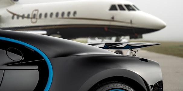 A luxury sports car with sleek black bodywork and blue accents is prominently featured in the foreground, showcasing its high-performance wheels. In the background, a private jet is parked on an overcast day, highlighting a scene of opulence and sophistication.