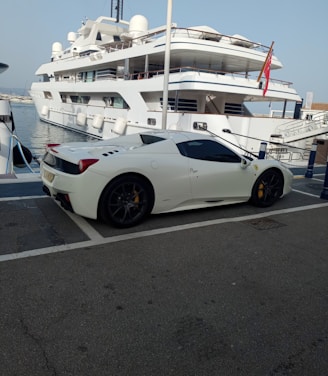 A luxury sports car parked near a large yacht, with both the car and yacht displaying sleek, modern designs. The setting appears to be a marina with calm water and other boats visible in the background.