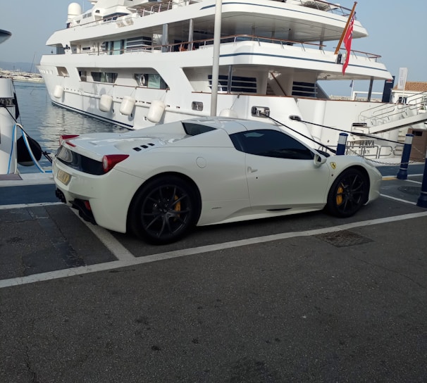 A luxury sports car parked near a large yacht, with both the car and yacht displaying sleek, modern designs. The setting appears to be a marina with calm water and other boats visible in the background.