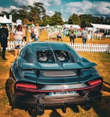 A luxury sports car is parked on a grassy area surrounded by people. The vehicle is sleek and modern with a shiny black exterior. In the background, a small crowd is gathered, with people standing near white picket fences and tents. The scene is set outdoors with trees and a partly cloudy sky.